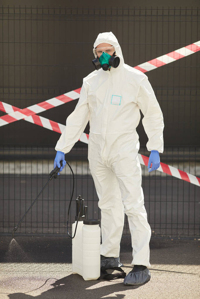 Vertikales Ganzkörperporträt eines männlichen Arbeiters im Warnanzug mit Desinfektionsausrüstung, während er im Freien steht, mit rotem Warnband im Hintergrund - Foto, Bild