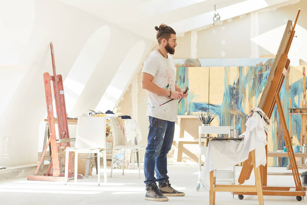 Full length portrait of contemporary male artist looking at painting while standing by easel in sunlit art studio, kopieer ruimte - Foto, afbeelding