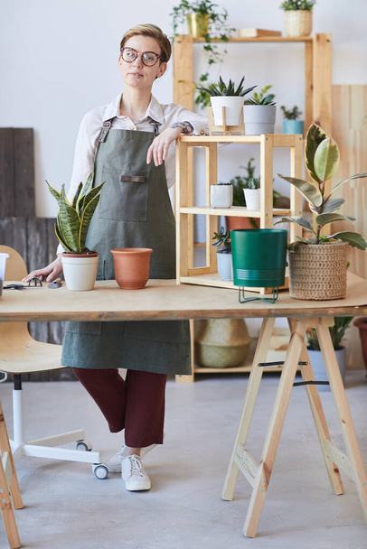 Retrato vertical de comprimento total da jovem mulher moderna vestindo avental em pé à mesa de madeira com plantas em vasos e solo pronto para o envasamento, conceito de jardinagem em casa
 - Foto, Imagem