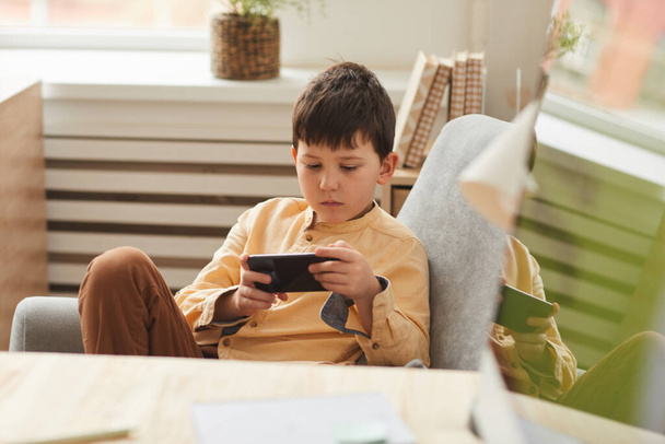 Koltuğunda oturan ve akıllı telefon ile mobil oyunlar oynayan sevimli bir çocuğun ılık tonlu portresi, boşluğu kopyala - Fotoğraf, Görsel