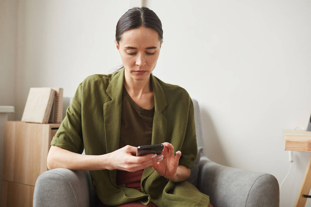 Portré a modern felnőtt nő használ okostelefon, miközben pihentető karosszék t otthon, másolás tér - Fotó, kép