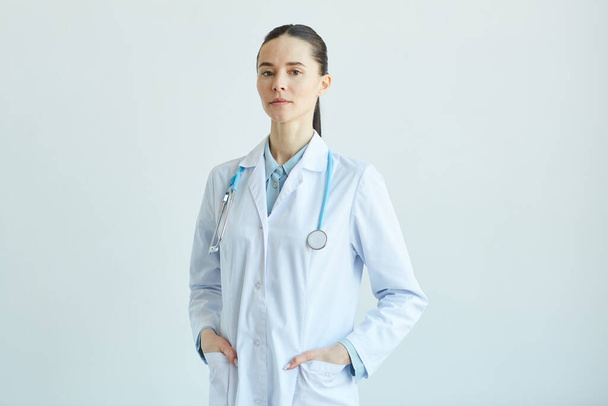 Waist up portrait of confident female doctor wearing lab coat and looking at camera while standing against white wall in clinic, copy space - Valokuva, kuva