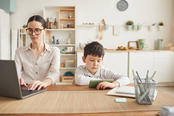 Portrait tonique chaud de la jeune mère travaillant au bureau à la maison avec son fils à l'aide d'un smartphone à côté d'elle dans un intérieur de cuisine confortable, espace de copie - Photo, image