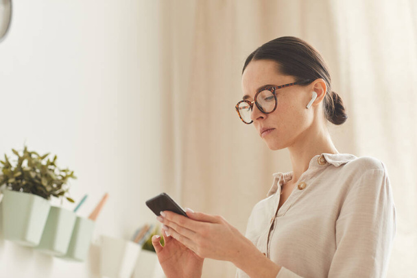 Retrato cálido de una mujer de negocios elegante usando un teléfono inteligente mientras está de pie en el acogedor interior de su casa, espacio para copiar - Foto, Imagen