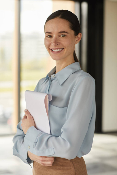 Vertical waist up portrait of elegant businesswoman smiling cheerfully and looking at camera while holding clipboard in empty office interior - Valokuva, kuva