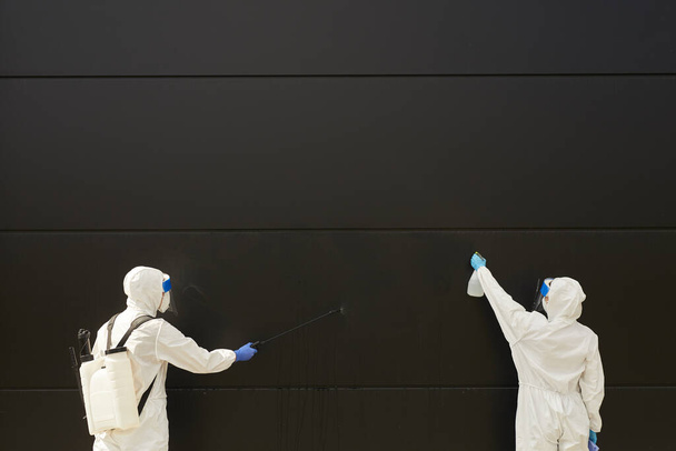 Vista grandangolare su due lavoratori che indossano indumenti protettivi e spruzzano sostanze chimiche sulla facciata nera dell'edificio durante la disinfezione o la pulizia, copiare lo spazio - Foto, immagini