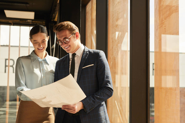 Waist up portrait of smiling rental agent showing floor plans to female client while standing in office building interior lit by sunlight, copy space - Foto, Imagen