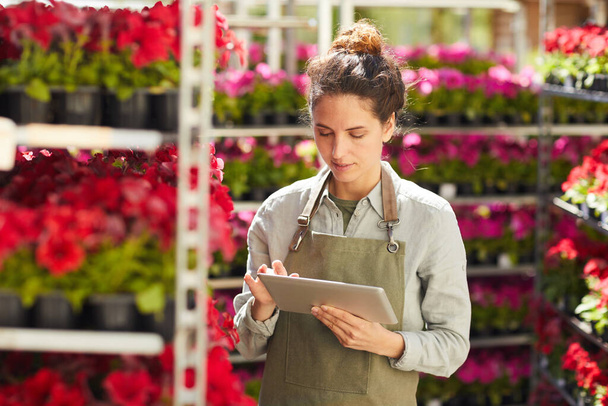 Retrato de cintura hacia arriba de una joven moderna usando tableta digital rodeada de hermosas flores mientras disfruta del trabajo en la plantación al aire libre, espacio para copiar - Foto, imagen