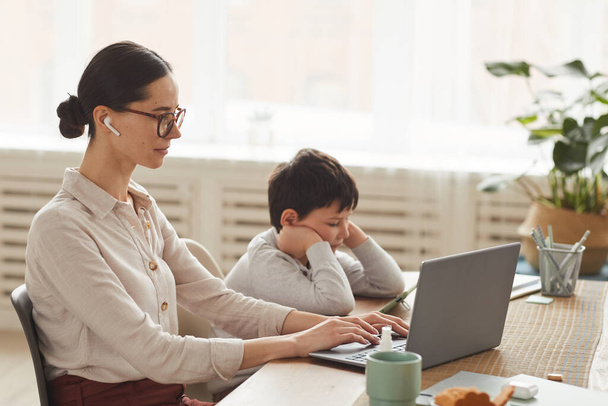 Warm getönte Seitenansicht Porträt einer jungen Mutter, die zu Hause im Büro arbeitet, während ihr Sohn das Smartphone neben sich in der gemütlichen Küche benutzt, Kopierraum - Foto, Bild