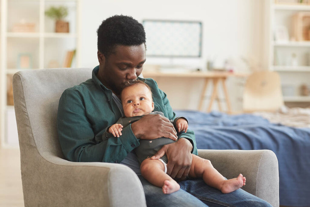Portrait of caring African-American man holding cute mixed-race baby while sitting in comfortable armchair at home, copy space - Фото, зображення