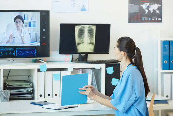 İş yerinde arka planda göğüs röntgeni görüntüleriyle otururken doktorla video görüşmesi yapan ve belgeleri gösteren genç kadın doktorların arka planı, boşluğu kopyala - Fotoğraf, Görsel