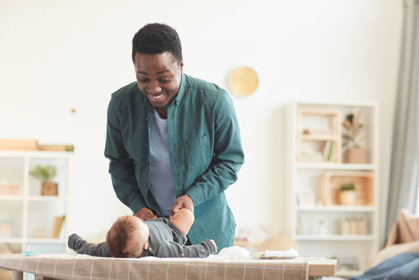 Retrato en tonos cálidos del amoroso padre afroamericano jugando con un lindo bebé mientras lo viste en el cambiador en casa, espacio para copiar - Foto, Imagen
