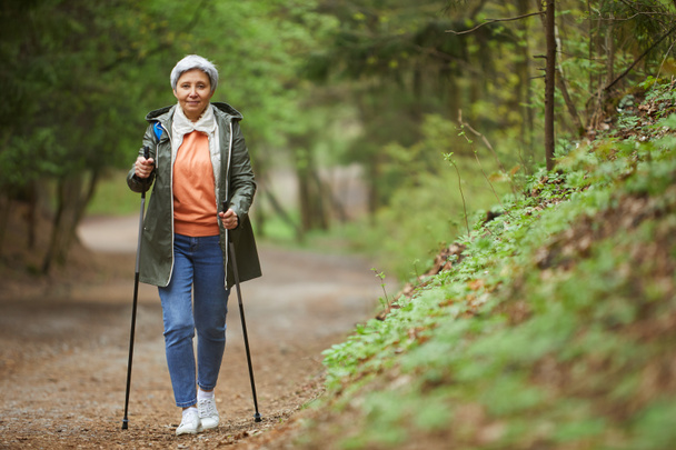 Full length portrait of active senior woman walking towards camera with Nordic poles while enjoying hike in beautiful autumn forest, copy space - Photo, Image