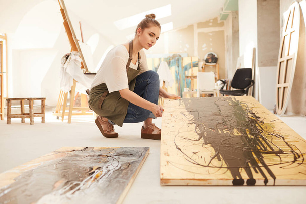 Warm-toned side view portrait of modern female artist holding abstract painting while sitting on floor in spacious art studio lit by sunlight, copy space - Φωτογραφία, εικόνα