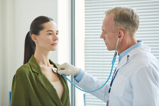 Side view portrait of mature doctor listening to breath and heartbeat of young woman via stethoscope while examining patient in clinic - Фото, изображение