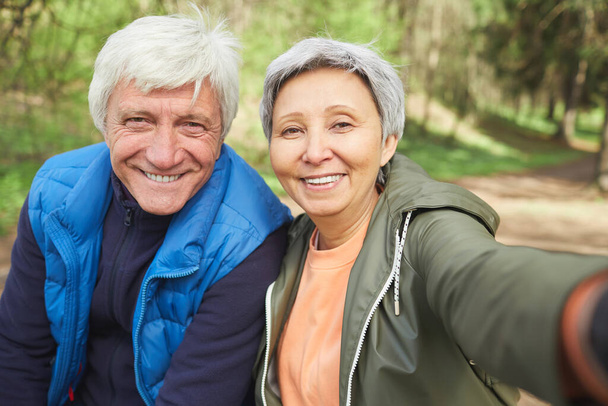 POV portrait of active senior couple looking at camera and smiling while taking selfie photo during hike in autumn forest, copy space - Photo, Image