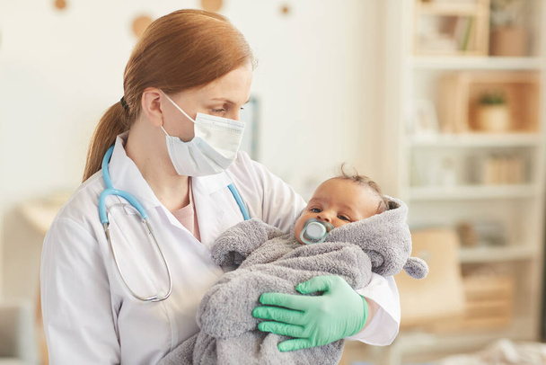 Retrato de cintura hacia arriba de una doctora sosteniendo un lindo bebé de raza mixta durante el chequeo en el interior del hogar, espacio para copiar - Foto, Imagen