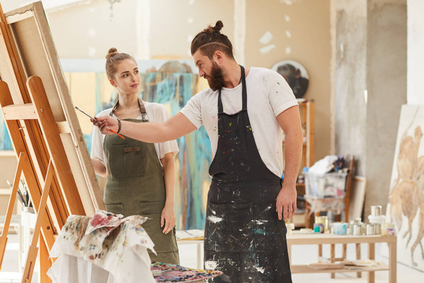 Retrato de pareja contemporánea pintando juntos mientras trabajan en el estudio de arte, se centran en el hombre barbudo que usa delantal y sostiene el pincel, espacio para copiar - Foto, Imagen