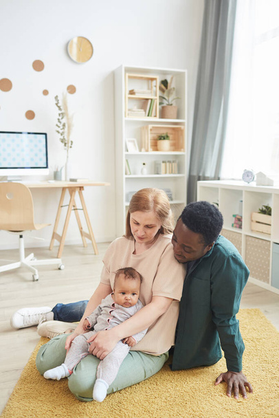 Pionowy portret pełnej długości współczesnej międzyrasowej rodziny bawiącej się słodkim mieszanym dzieckiem podczas siedzenia na podłodze w domu, przestrzeń kopiowania - Zdjęcie, obraz