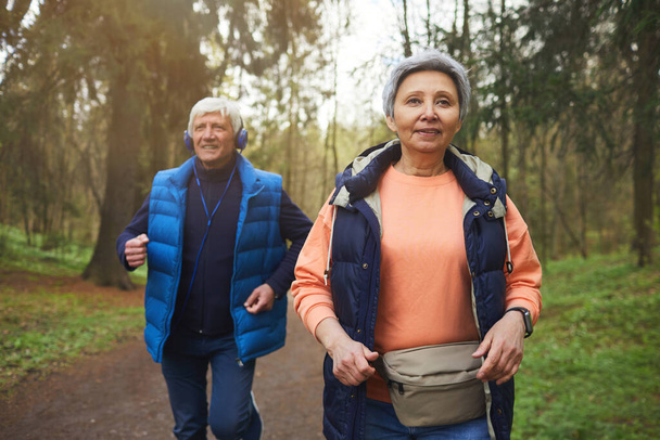 Pasie w górę portret aktywnej pary seniorów biegnącej w kierunku kamery w pięknym jesiennym lesie i uśmiechającej się szczęśliwie - Zdjęcie, obraz