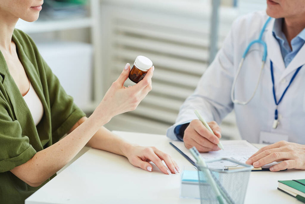 Mid-section of unrecognizable female patient holding bottle of pills while sitting at desk in doctors office during consultation, copy space - Photo, image