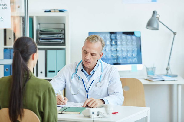 Portrait of mature male doctor writing on clipboard while sitting at desk and consulting female patient in modern clinic, copy space - Foto, Imagen