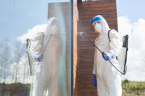Πλευρική άποψη πορτρέτο του άνδρα εργαζομένου που φοράει προστατευτική στολή ψεκάζοντας χημικές ουσίες πάνω από το κτίριο σε εξωτερικούς χώρους κατά την απολύμανση, αντίγραφο χώρου με αντανάκλαση - Φωτογραφία, εικόνα