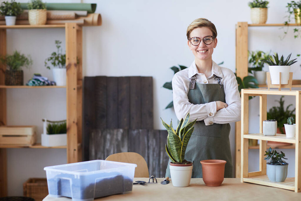 Талия портрет улыбающейся молодой женщины в фартуке, стоящей у деревянного стола с горшечными растениями и почвой, готовой к горшку, домашний садоводческий концепт, копировальное пространство - Фото, изображение