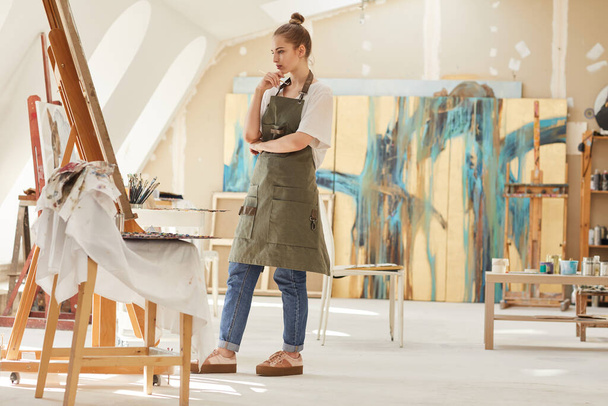 Portrait complet d'une artiste féminine moderne regardant la peinture tout en travaillant dans un atelier d'art éclairé par la lumière du soleil, espace de copie - Photo, image