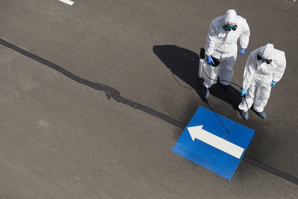 Above view at two workers wearing protective gear and spraying chemicals outdoors standing on concrete road with blue arrow pointing left, copy space - Fotoğraf, Görsel