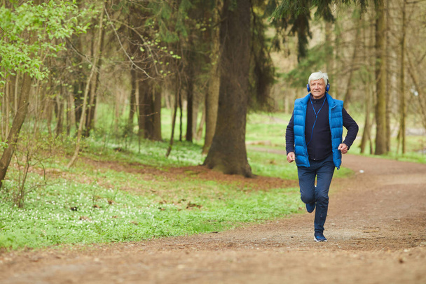 Širokoúhlý portrét aktivního staršího muže poslouchajícího hudbu při běhu v krásném podzimním lese, kopírovací prostor - Fotografie, Obrázek