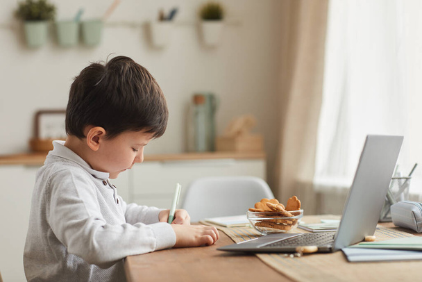 Lämmin sävyinen sivunäkymä muotokuva söpö poika tekee kotitehtäviä, kun istuu pöydällä kodikas sisustus, kopioi tilaa - Valokuva, kuva