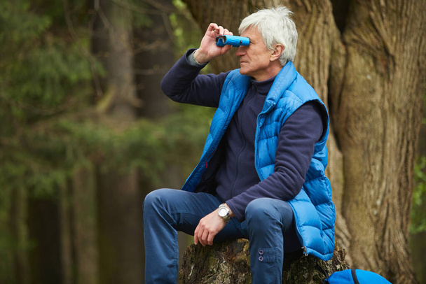 Portrait of active senior man looking in binoculars while sitting by tree during hike in autumn forest, copy space - Photo, Image