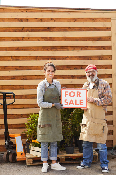 Vertikales Ganzkörperporträt zweier zeitgenössischer Gärtner, die ein rotes zum Verkauf stehendes Schild halten, während sie im Freien vor einer Plantage stehen, Kopierraum - Foto, Bild
