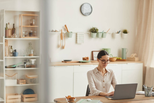 Теплый широкоугольный портрет элегантной молодой женщины в очках во время использования ноутбука на уютном домашнем рабочем месте, копировальное пространство - Фото, изображение