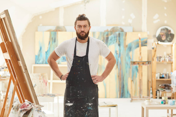 Portrait of mature bearded artist wearing apron and posing confidently looking at camera while standing in spacious art studio lit by sunlight, copy space - Photo, Image