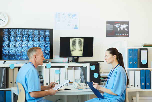 Widok z boku portret dwóch dorosłych lekarzy mówiących podczas siedzenia w miejscu pracy we wnętrzu nowoczesnej kliniki z tomografią komputerową w tle, przestrzeń do kopiowania - Zdjęcie, obraz