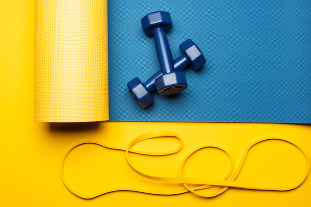 vista dall'alto del tappeto fitness blu con manubri e banda di resistenza su sfondo giallo - Foto, immagini