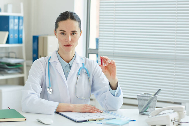 Portret profesjonalnej lekarki trzymającej próbkę krwi i patrzącej w kamerę, siedzącej przy biurku w nowoczesnej klinice, przestrzeń do kopiowania - Zdjęcie, obraz