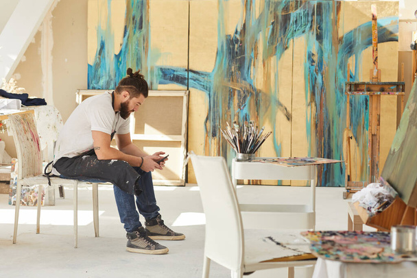 Vue latérale sur toute la longueur de l'artiste barbu contemporain en utilisant un smartphone assis sur une chaise dans un studio d'art avec des peintures abstraites en arrière-plan, espace de copie - Photo, image