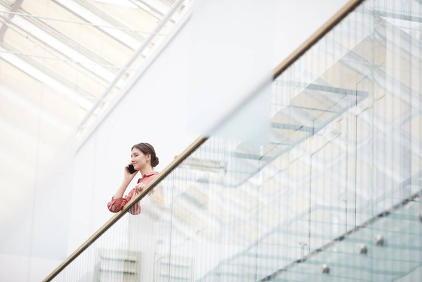 Широкий угол обзора на молодую деловую женщину, говорящую по смартфону, стоя на балконе в минимальном интерьере офисного здания, копировальное пространство - Фото, изображение