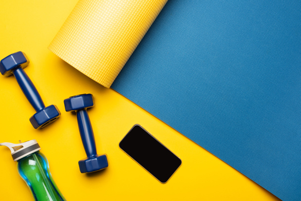vue du dessus du tapis de fitness bleu avec haltères, smartphone et bouteille de sport sur fond jaune - Photo, image