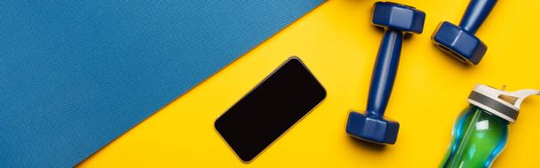 vrchní pohled na modrou fitness podložku s činkami, smartphone a sportovní láhev na žlutém pozadí, panoramatický záběr - Fotografie, Obrázek