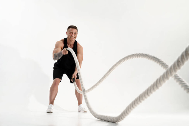 Attractive muscular man working out with heavy ropes. Photo of handsome man in sportswear isolated on white background - Foto, Imagen