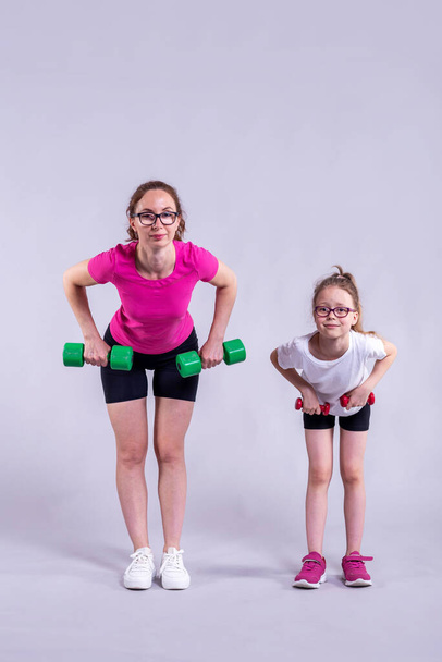 Stylish mom and daughter stand on a gray background in sportswear and with dumbbells. Active lifestyle, fitness. Sport family. - Фото, изображение