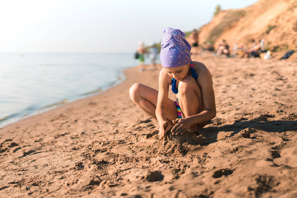 Mayolu ve bandanalı küçük bir kız çocuğu onu güneşten koruyor kumsalda kumsalda oturuyor ve kumlarla oynuyor, kaleler inşa ediyor, yazı yazabileceği bir yer, yaz tatili konsepti. - Fotoğraf, Görsel