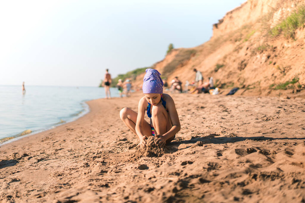 A little girl child in a swimsuit and a bandana protecting her from the sun sits on the sand on the beach and plays with the sand, builds castles, a place for text, the concept of a summer vacation - Photo, Image