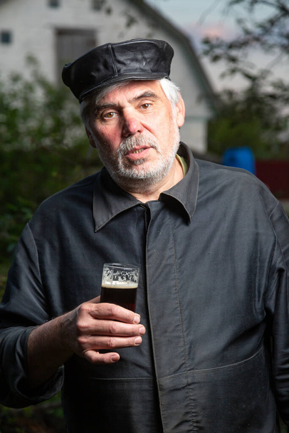 Serious kind sad elderly man with black old-fashioned old clothes and a leather cap with a glass of beer in his hand. Looks at the camera. Vertical orientation. - Photo, Image