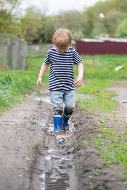 a small boy in rubber boots walks through a muddy puddle - Foto, afbeelding