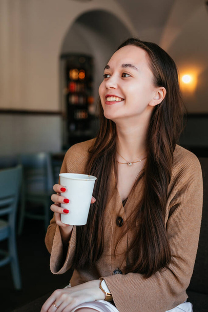 Close portrait of an attractive beautiful girl holding and drinking a cup of coffee in her hand and smiling. Positive student girl resting in a cafe, drinking coffee in a coffee shop - Photo, image